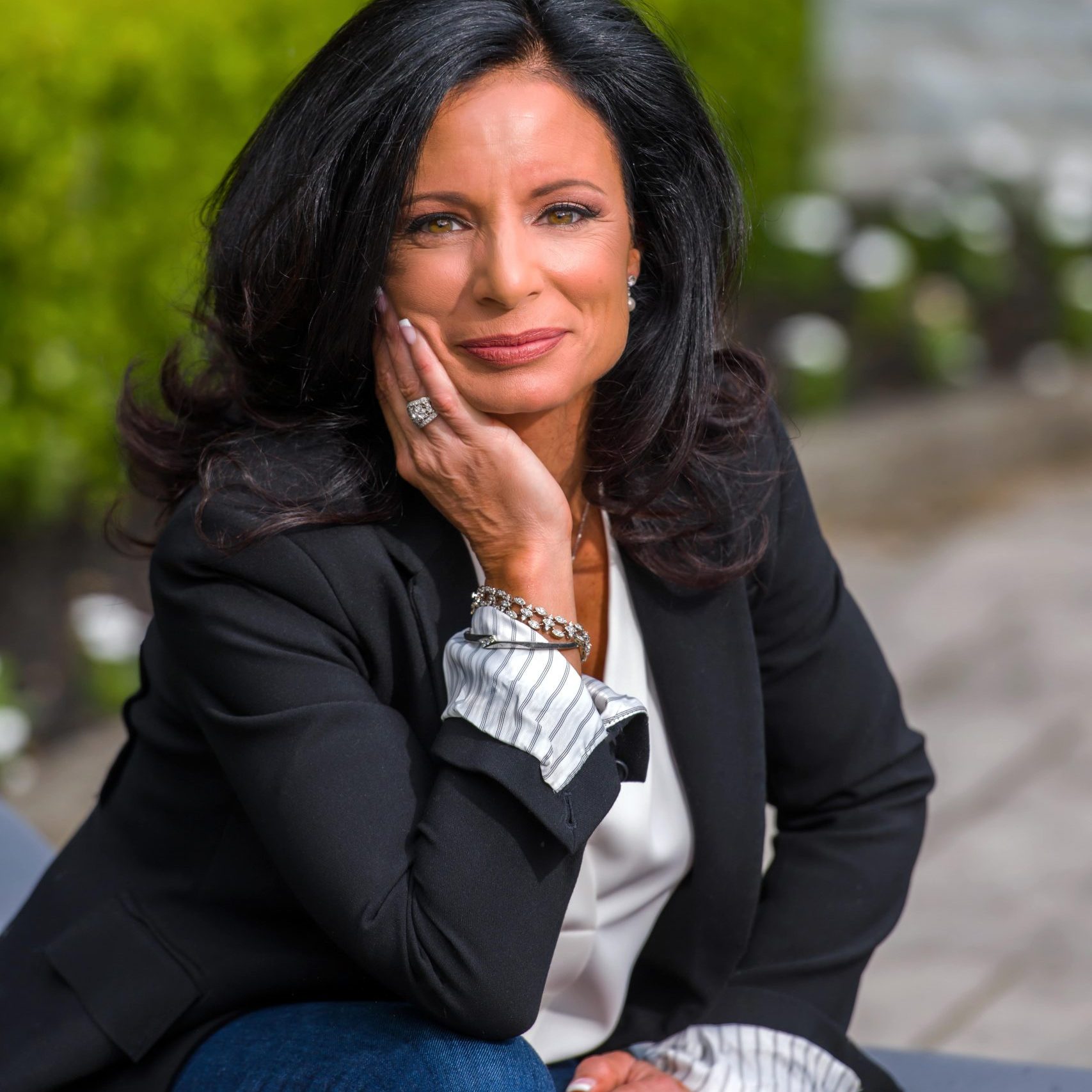 Image of a thoughtful woman with dark hair, dressed in a smart black blazer and blue jeans, sitting outdoors with green hedges in the background, resting her head on her hand.